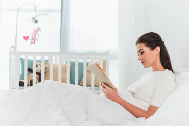側ビューの女性読書本上のベッドに近いぼやけた赤ちゃんクリブ  - 写真・画像
