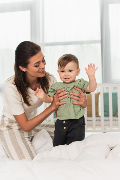 Cheerful woman holding son waving hand on bed  - Foto, Imagen