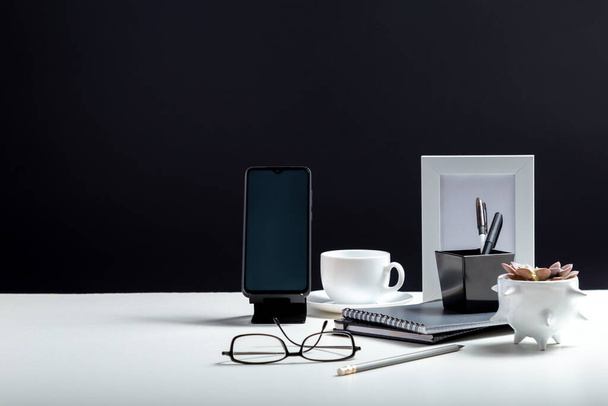 Office or home Office workspace. Desktop with smart phone template blank, notepads pens office suppliers cup of coffee drink plant flower. White work desk table on black background with copy space. - Fotoğraf, Görsel