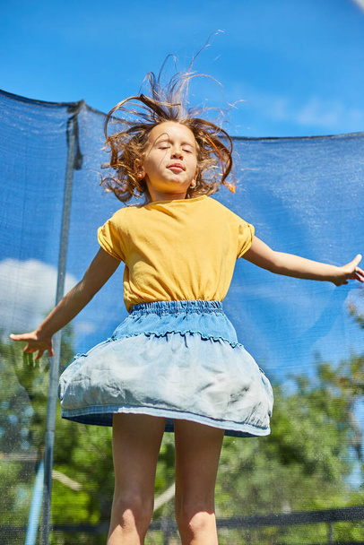 Een jong meisje springen op en neer op haar trampoline buiten, in de achtertuin van het huis op een zonnige zomerdag, zomervakantie. - Foto, afbeelding