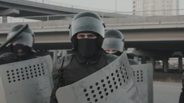 Slowmo tracking shot of riot police officer in gear holding shield and baton and walking up to pose before camera while other policemen standing in formation behind him - Footage, Video