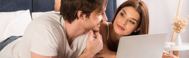 happy couple looking at each other while watching film on laptop in bedroom, banner - Photo, Image
