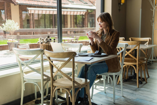 Freelancer κρατώντας κύπελλο κοντά επιδόρπιο και φορητό υπολογιστή στο καφέ  - Φωτογραφία, εικόνα