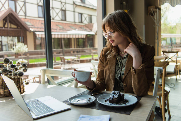Stylish woman with cup sitting near chocolate dessert and laptop in cafe - Foto, Bild