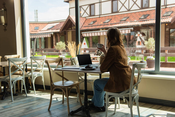 Stylish woman holding cup near delicious dessert and laptop in cafe  - Fotografie, Obrázek
