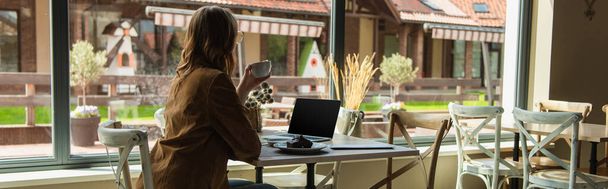 Young woman with cup looking at window near cake and laptop in cafe, banner  - Фото, изображение