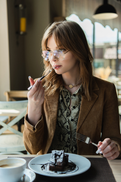 Μοντέρνα γυναίκα με γυαλιά που τρώει επιδόρπιο κοντά στο θολό καφέ στο καφέ  - Φωτογραφία, εικόνα