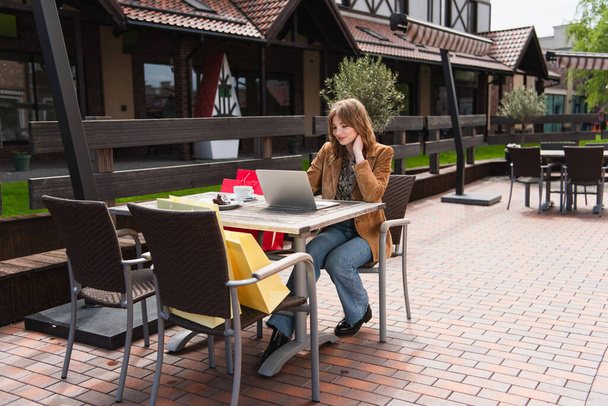 Woman using laptop near cup, dessert and shopping bags on terrace  - Photo, Image