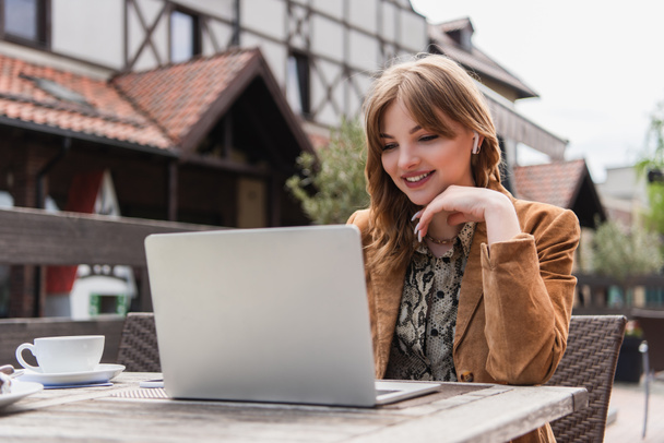 Freelance souriant dans l'écouteur en utilisant un ordinateur portable près du café sur la terrasse - Photo, image