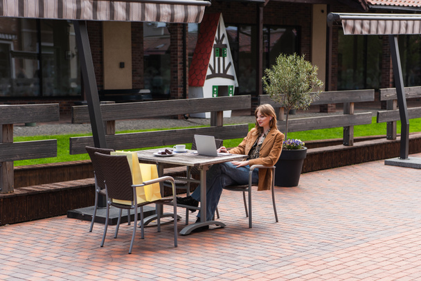 Freelancer de moda con portátil cerca de café, postre y bolsas de compras en la terraza de la cafetería  - Foto, imagen