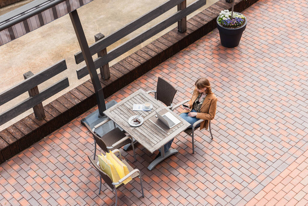 Overhead view of woman using smartphone near laptop, coffee and purchases on terrace  - Φωτογραφία, εικόνα