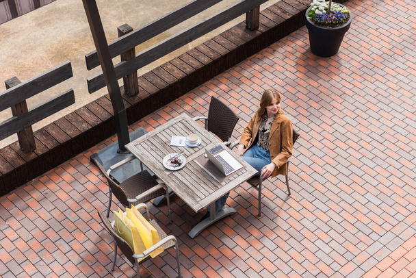 Blick von oben auf stilvolle Frau, die in der Nähe von Gadgets, Dessert und Einkaufstaschen auf der Terrasse sitzt  - Foto, Bild