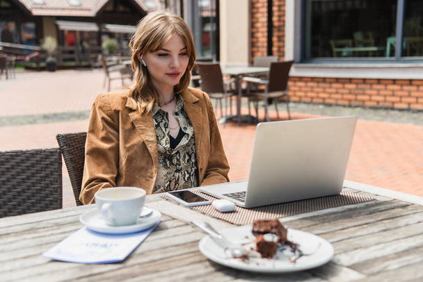 Νεαρή γυναίκα στο ακουστικό κοιτάζοντας το φορητό υπολογιστή κοντά στον καφέ και θολή τούρτα στη βεράντα του καφέ  - Φωτογραφία, εικόνα