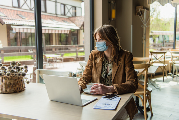 Woman in medical mask sitting near gadgets and menu in cafe  - Fotoğraf, Görsel