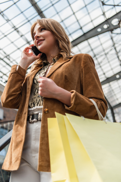 Tiefansicht einer stilvollen Frau mit verschwommenen Einkaufstüten, die in einem Einkaufszentrum auf dem Handy spricht  - Foto, Bild