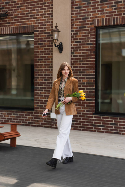 stylová žena s chytrým telefonem drží tulipány a chůze venku - Fotografie, Obrázek