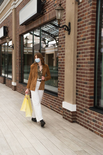 full length of young stylish woman in medical mask walking with shopping bags near building of mall - Zdjęcie, obraz
