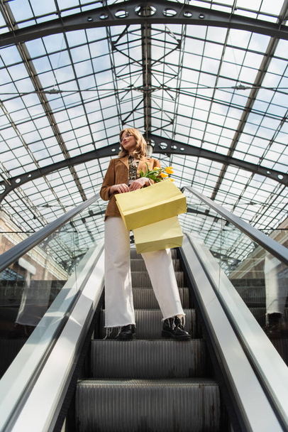 teljes hossza nő napszemüvegben tartó sárga tulipán és bevásárló táskák, miközben álló mozgólépcső  - Fotó, kép