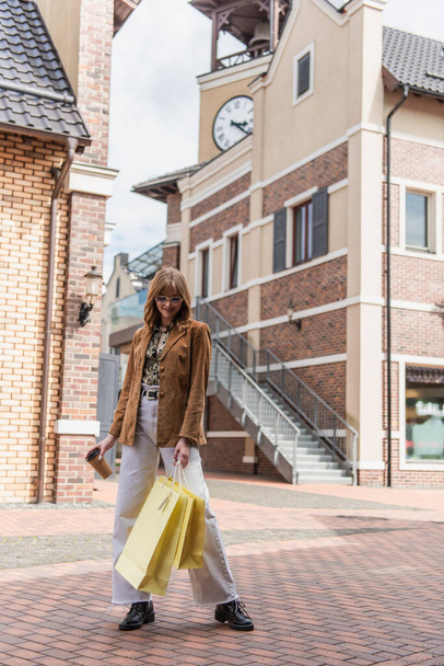 full length of cheerful woman in sunglasses and jacket holding shopping bags and paper cup  - Foto, imagen