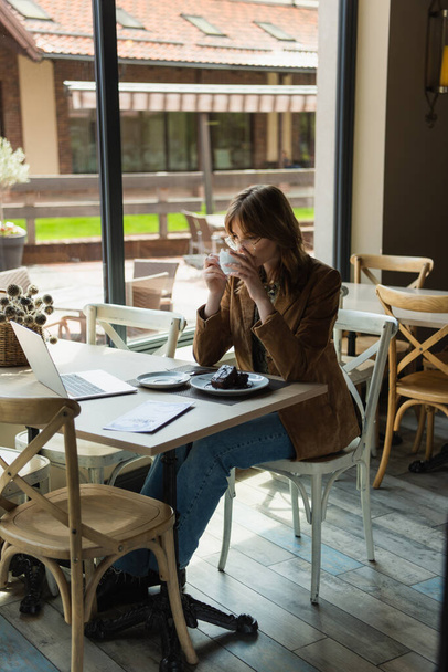 junge Frau mit Brille blickt auf Laptop in der Nähe von Schokoladenkuchen und trinkt Kaffee im Café - Foto, Bild