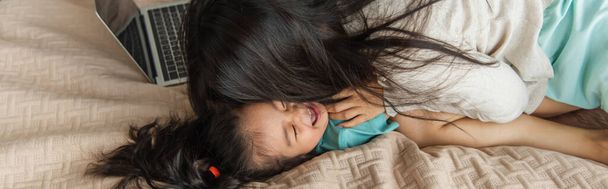 Мать целует азиатского ребенка возле ноутбука на кровати, баннер  - Фото, изображение