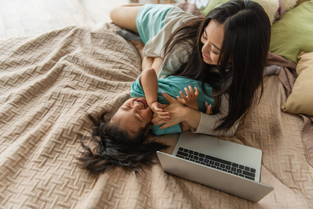 Vue aérienne de la mère s'amuser avec un enfant asiatique près d'un ordinateur portable sur le lit  - Photo, image