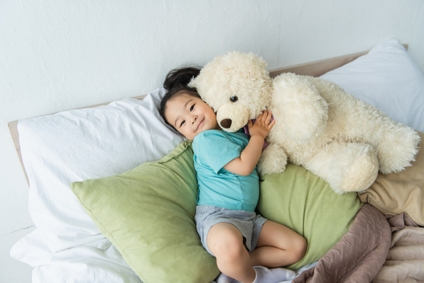 sonriente asiático niño holding teddy oso en cama  - Foto, Imagen