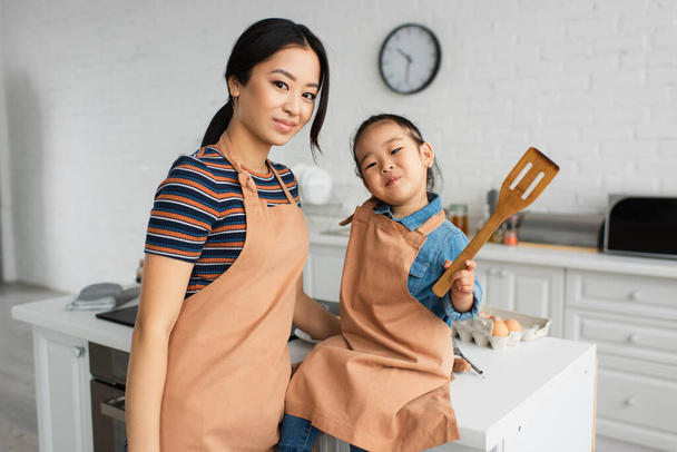 Asian woman looking at camera near daughter with spatula in kitchen  - Fotoğraf, Görsel