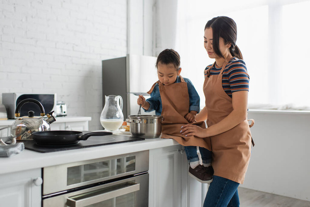 Smiling asian woman standing near daughter with whisk in kitchen  - Photo, image