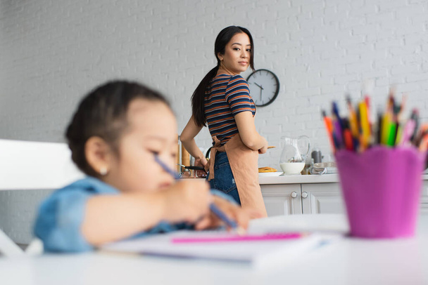 Asian woman looking at daughter drawing while cooking in kitchen  - Foto, afbeelding