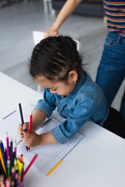 Asian girl drawing near blurred color pencils and parent at home  - Photo, Image
