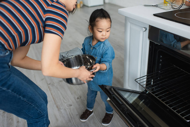 Asian kid in oven glove holding pan near mother and oven  - Photo, image