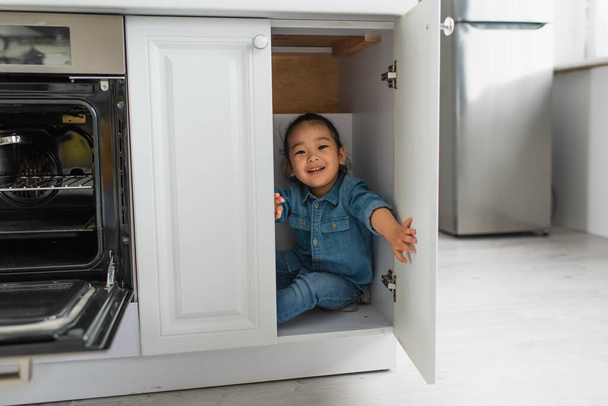 Улыбающаяся азиатка смотрит в камеру из кухонного шкафа  - Фото, изображение