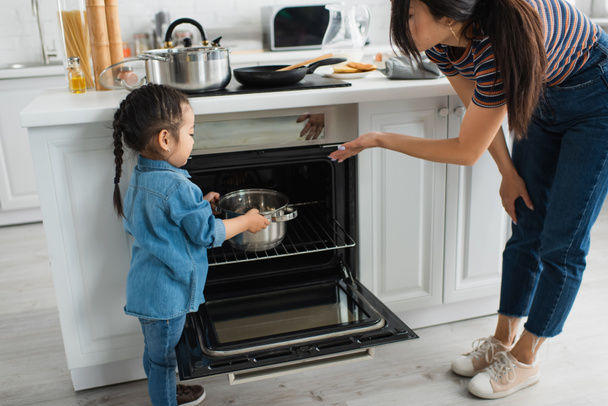 Mother pointing at oven near asian kid with pan  - Foto, imagen