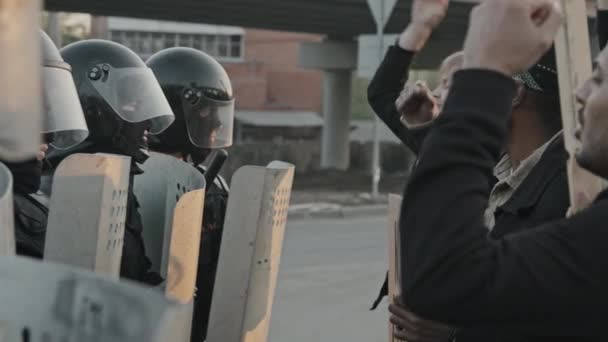 Langsam aufgenommene Aufnahmen von Bereitschaftspolizisten in voller Ausrüstung, die Schilde halten und in Reihe stehen, während junge Leute singen und protestieren - Filmmaterial, Video