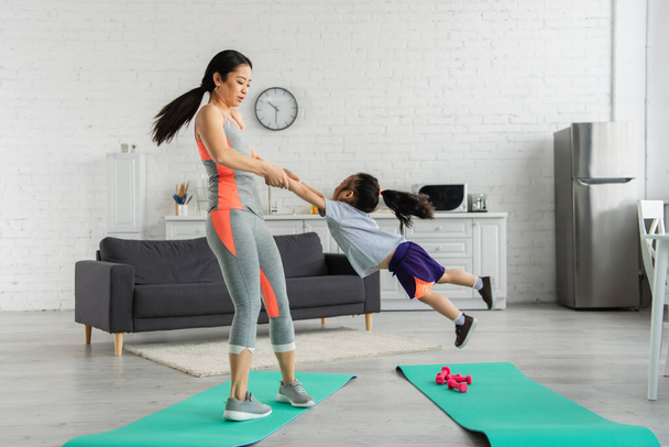 Mulher asiática girando filha perto de tapetes de fitness em casa  - Foto, Imagem