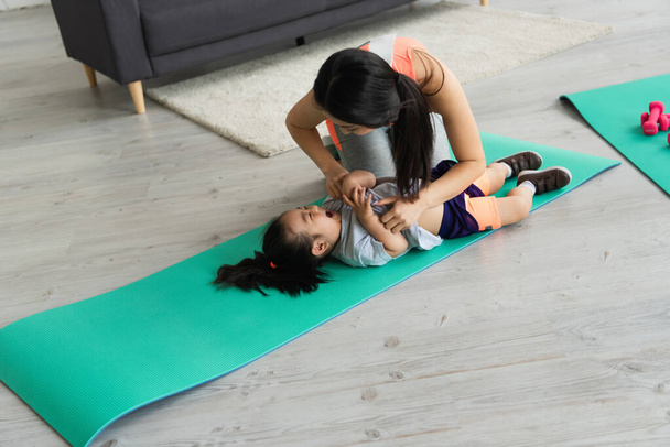 Mother tickling cheerful asian kid on fitness mat  - 写真・画像