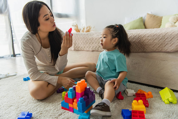 Asian mother looking at building block near daughter on carpet  - Foto, afbeelding