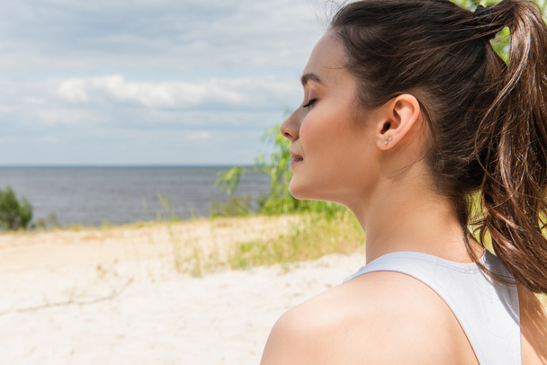 young and brunette woman with closed eyes meditating near ocean - Photo, Image