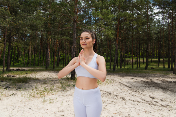 smiling young woman in crop top and leggings standing with praying hands in forest - Photo, Image
