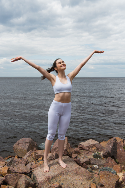 szczęśliwa młoda kobieta z zamkniętymi oczami i podniesionymi rękami w pobliżu morza  - Zdjęcie, obraz