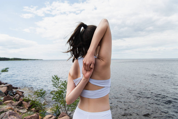 back view of young woman doing cow face pose with hands behind back near sea - Valokuva, kuva