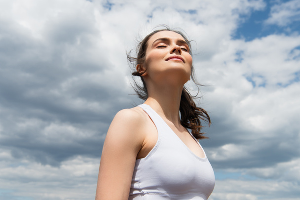 visão de ângulo baixo de mulher jovem com olhos fechados contra o céu azul com nuvens  - Foto, Imagem