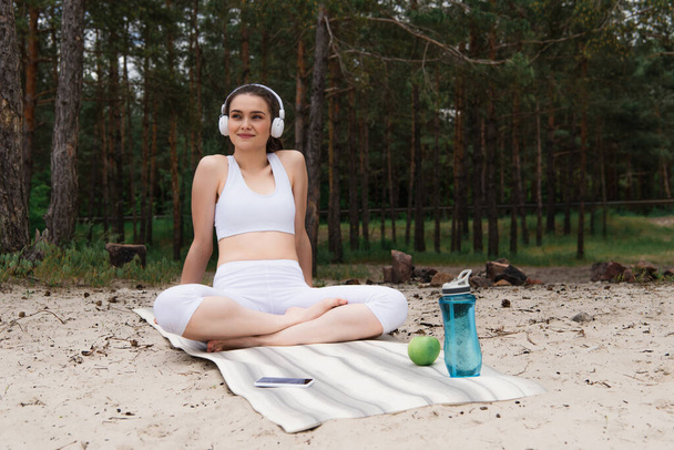 happy woman in headphones listening music while sitting with crossed legs on yoga mat  - Photo, Image