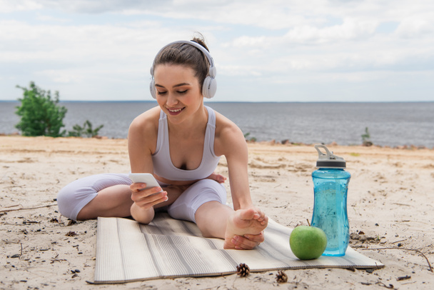 улыбающаяся женщина в наушниках слушает музыку, растягиваясь на коврике для йоги и используя смартфон  - Фото, изображение