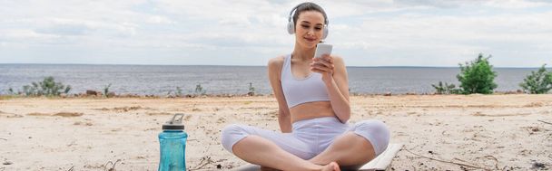 uśmiechnięta kobieta w słuchawkach słuchająca muzyki podczas korzystania ze smartfona i siedząca ze skrzyżowanymi nogami na macie do jogi, baner - Zdjęcie, obraz