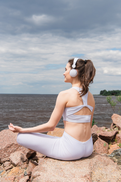 mujer feliz en auriculares y ropa deportiva sentado en pose de loto mientras medita cerca del mar  - Foto, imagen