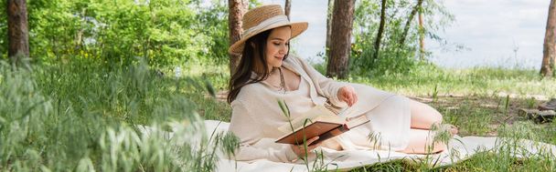 χαρούμενη νεαρή γυναίκα με ψάθινο καπέλο διαβάζει βιβλίο στο δάσος, πανό - Φωτογραφία, εικόνα