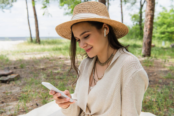 positive young woman in straw hat and wireless earphones using smartphone in forest  - Fotografie, Obrázek