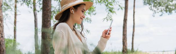 positive young woman in straw hat and wireless earphones using smartphone in forest, banner - Φωτογραφία, εικόνα